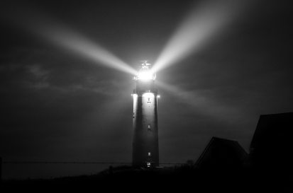 A gray scale photo of a lighthouse at night, with light shining in all directions.
