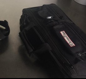 A black messenger bag and a black knife roll on a steel table with knives in the background.