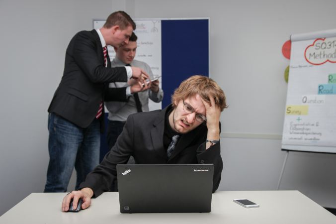 stressed out man grimacing at a black laptop while two other men talk behind him