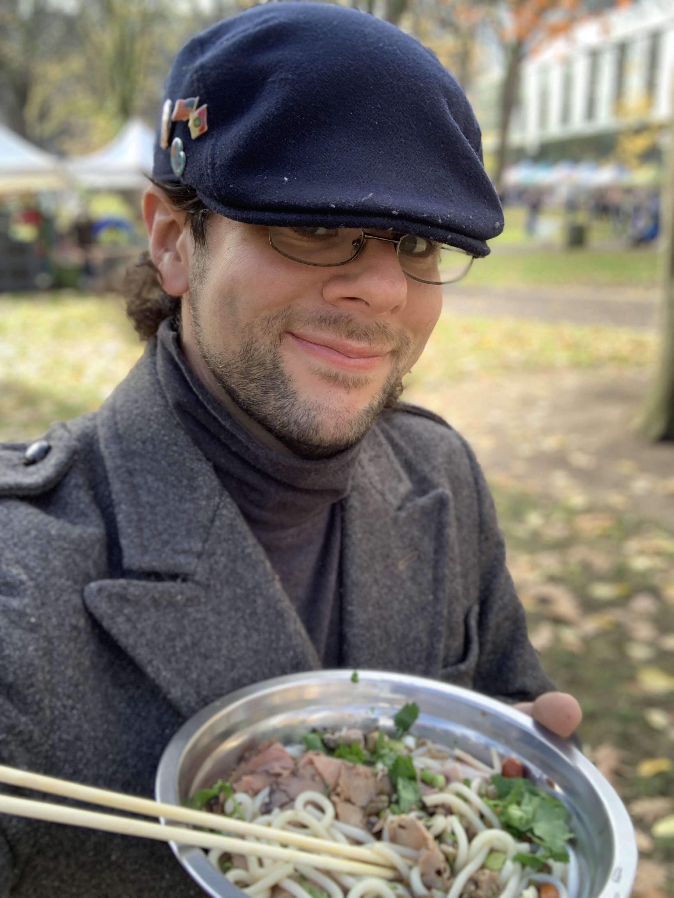 Selfie of the author in a gray wool coat and blue cap, holding up a bowl of noodle soup with chopsticks. It is outside on a fall day.