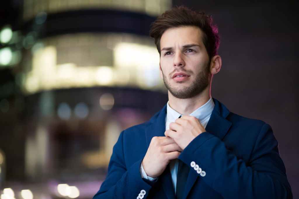 A young man with a thin beard and a blue business suit adjusting his necktie