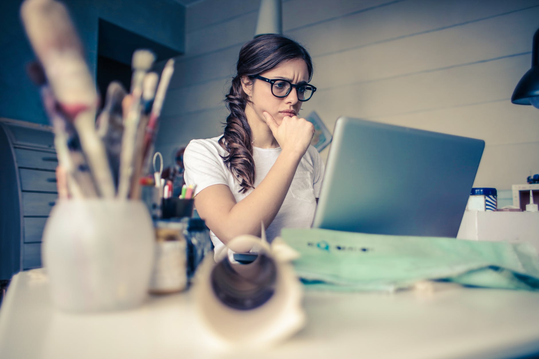A young woman with glasses and a cluttered desk reads a computer screen intently