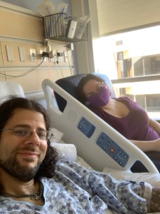 Selfie of the author and his wife. Matt is in a hospital bed and gown, his wife sitting beside him in a chair with  mask on.