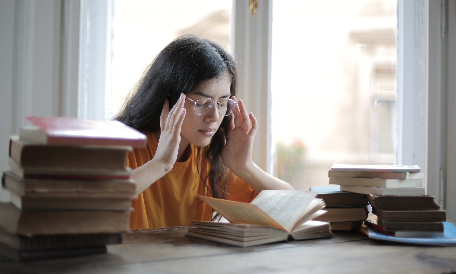 a young woman with long hair presses her fingers to her temples surrounded by books at a library table