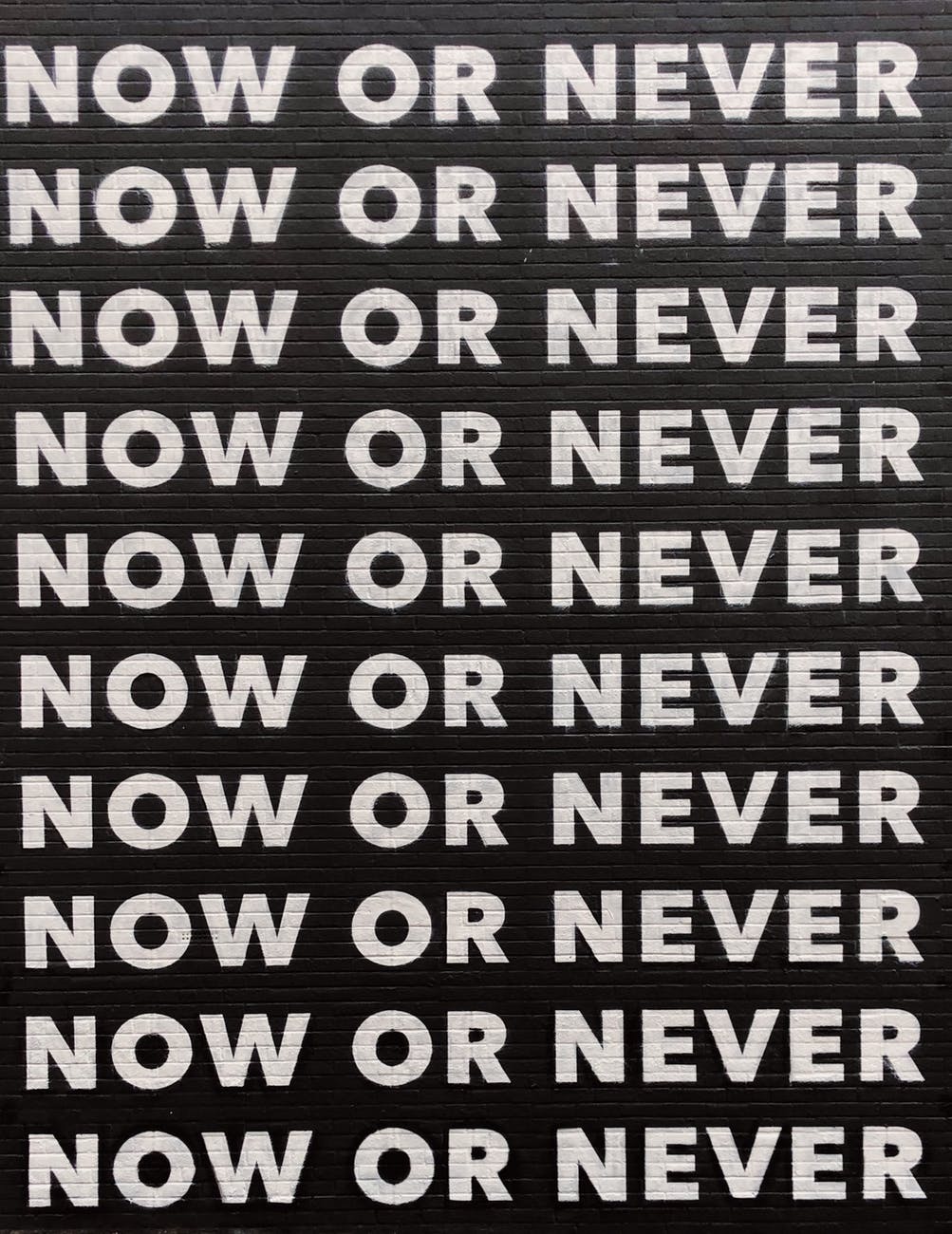 Repeated white text on a black background that looks like brick: "Now or Never."