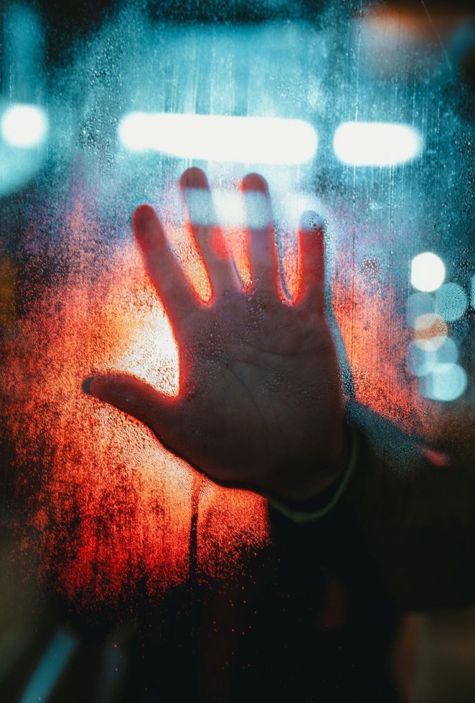 A hand pressed against fogged, wet glass. White and red lights of an ambulance shine through from the other side.