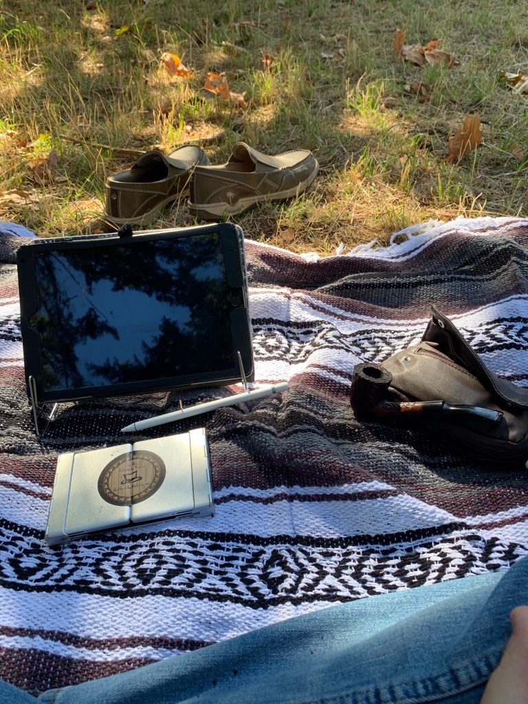 A striped Mexican blanket is on a grassy field. On the blanks are a pair of loafers, an ipad, a folding keyboard, and a tobacco pipe.