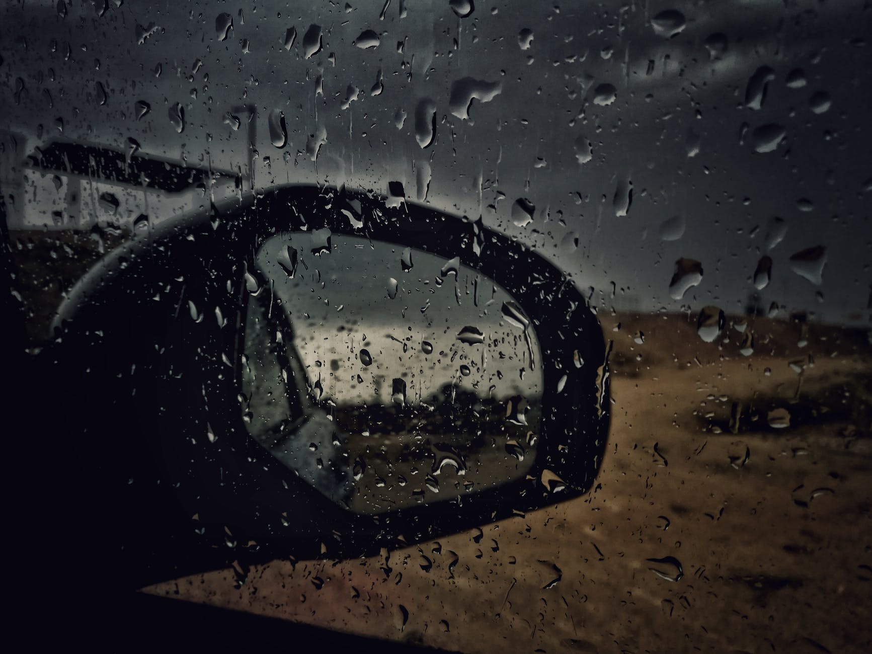 Rain on a car window looking at the side-view mirror down an empty road.