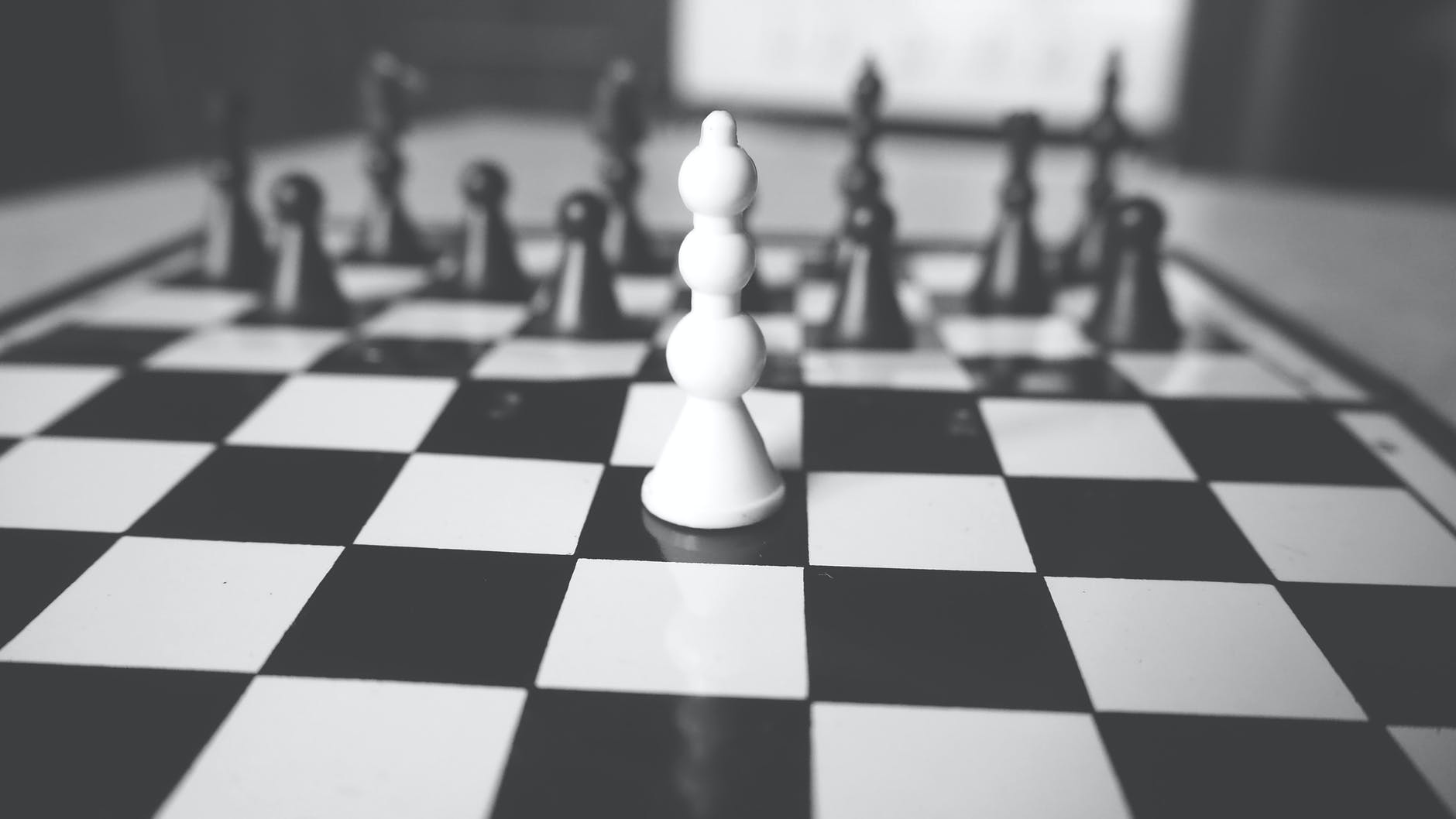 black and white picture of a chessboard, with a single white bishop in front of a full black setup.