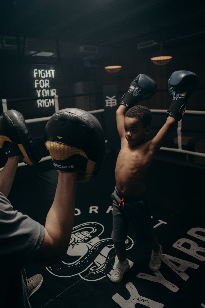A young black boy in boxing gloves raises in arms in victory in front of his trainer who is wearing punching pads.
