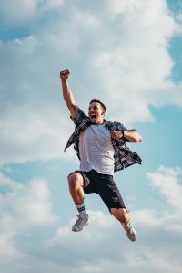 A young man in a flannel shirt, white tshirt and shorts jumping in the air against a blue sky and pumping his fist.