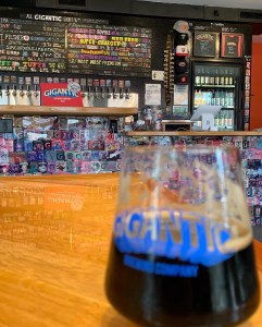 Taken Gigantic Brewings “Robot Room”- a half-full glass of beer is unfocused in the foreground while the bar and it’s extensive tap list are focused in the back. The bar is covered in bright colors and cartoonish labels.