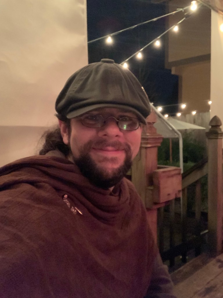 The author sits on a dimly lit patio, smiling. He is wrapped in a brown shawl.