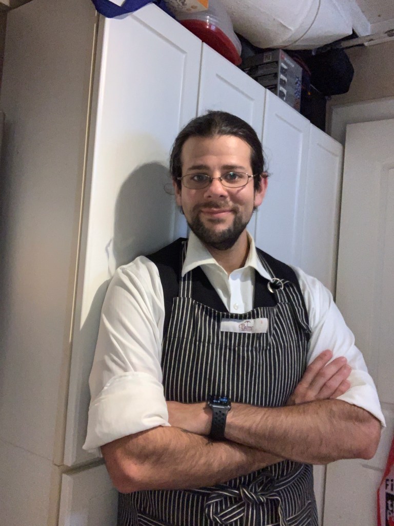 The author is wearing an apron over a black suit vest and white button-front shirt with the collar open and sleeve rolled up. He's smiling at the camera and leaning against white cabinets.