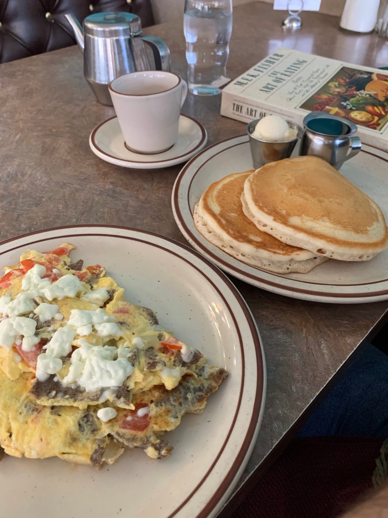 A breakfast on a diner table. An omelette with feta cheese is on top in the foreground. Behind it is a plate with two fluffy pancakes and cups of butter and syrup. Behind that a cup of tea, and a copy of “The Art of Eating” by M.F.K. Fisher.