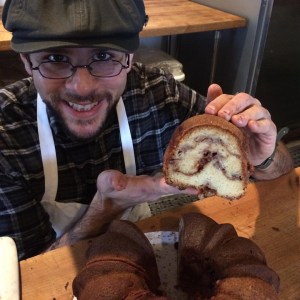 A young man with a goatee smiles and proudly holds up a cross-section of a Jewish Apple Bundt Cake.