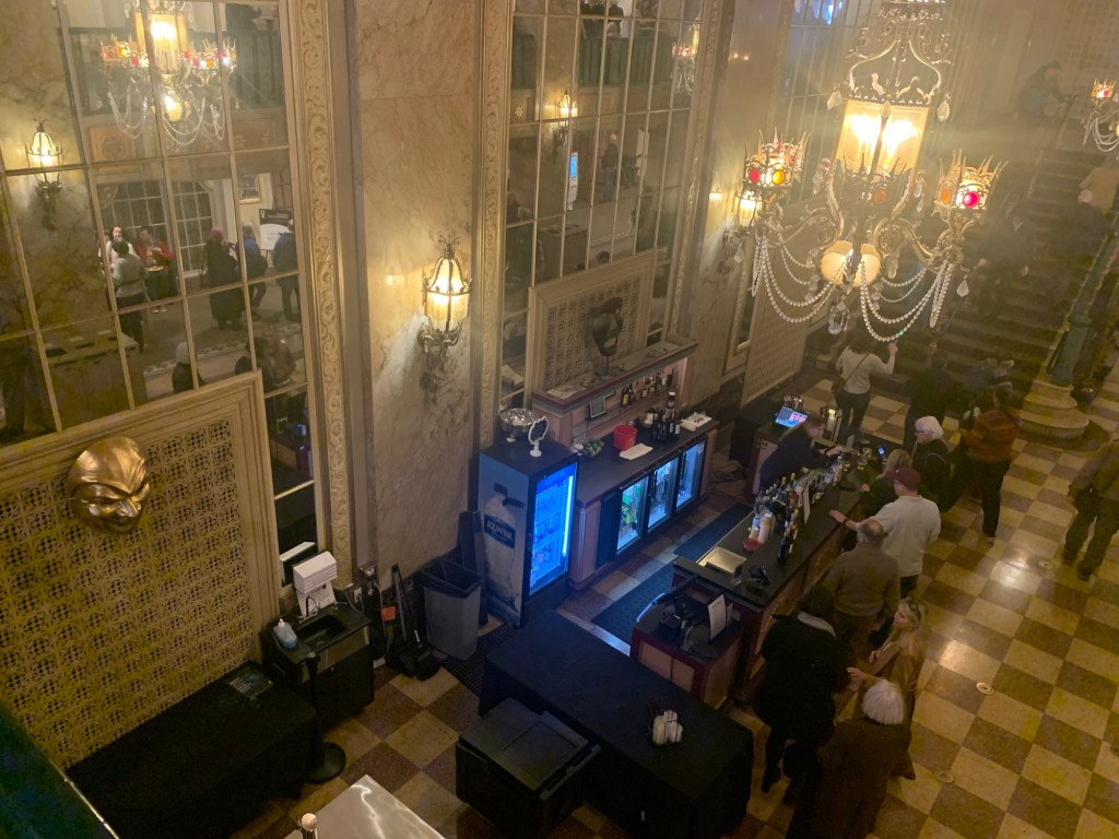 View of a refreshment bar in the lobby of a theater, taken from a balcony looking down. There are soda coolers and a limited liquor selection, overseen by two large golden theater masks.