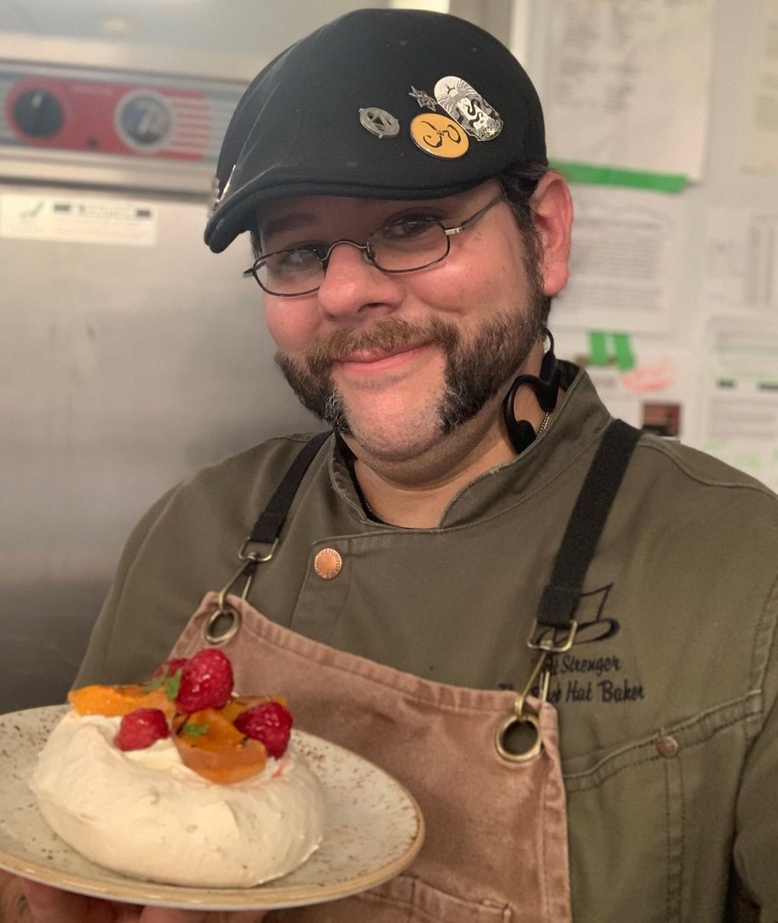 A selfie of the author- black flat-cap with pins, olive green chef coat, brown canvas apron, smiling face with mutton-chops, and holding up a plated dessert