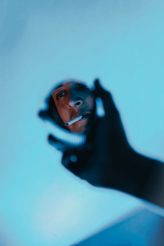 The silhouette of a hand holding a small mirror. In the mirror is a man- apparently the one holding the mirror- with a cigarette in his mouth and looking into the distance.